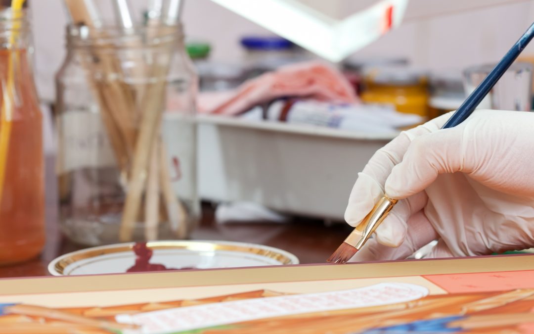 A gloved hand uses a paintbrush to work on a colorful canvas, with art supplies in the background.