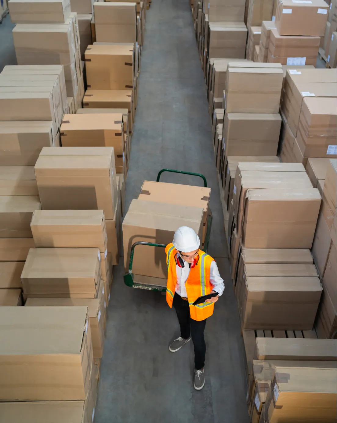 Worker in safety vest and helmet uses a tablet in a warehouse surrounded by stacks of cardboard boxes.