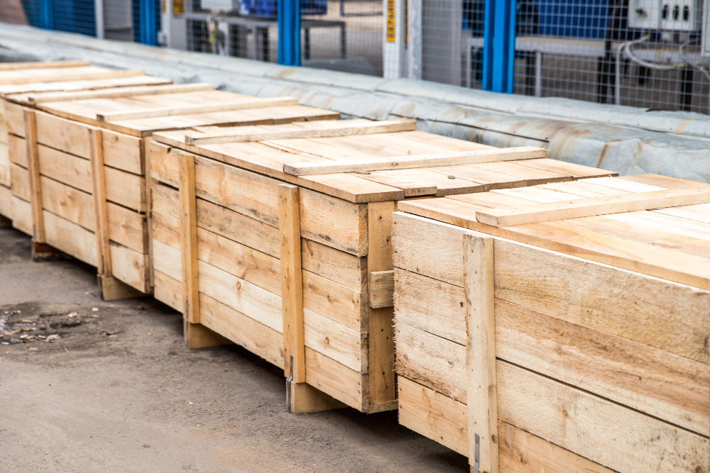 Several large wooden crates are lined up outdoors near some metal fencing and industrial equipment.